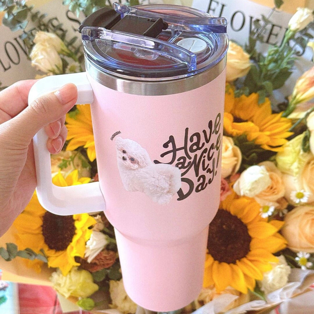 Pink tumbler with 'Have a Nice Day' text and cartoon character, held in front of sunflower bouquet.
